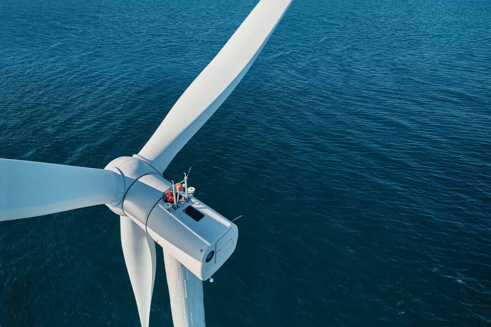 Aerial view of a wind turbine above blue ocean water, with a technician working on the turbine's hub.
