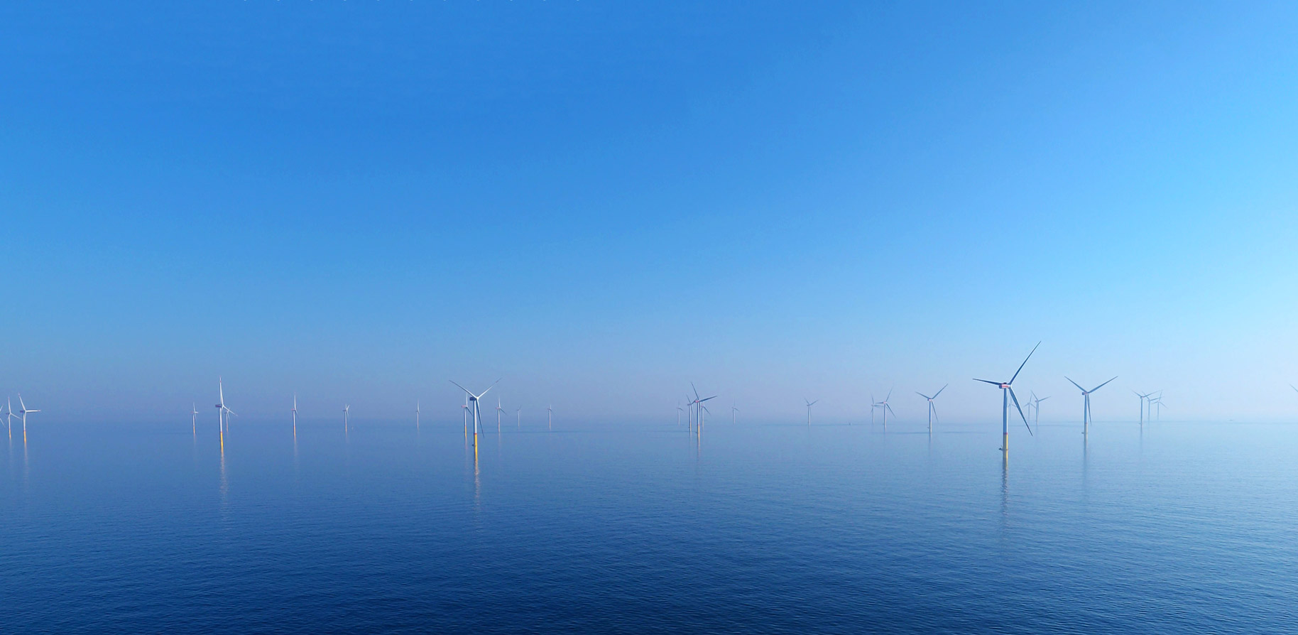 Een heldere blauwe lucht boven een rustige zee, bezaaid met meerdere windturbines die offshore draaien.