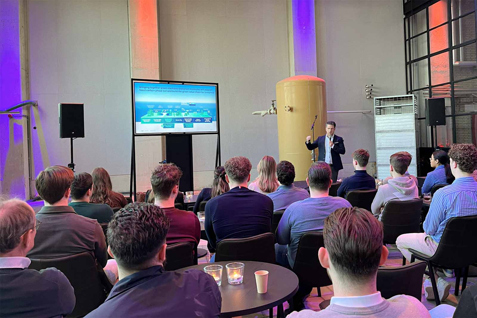 A speaker presents a digital slide in a modern venue to an engaged audience seated at tables.