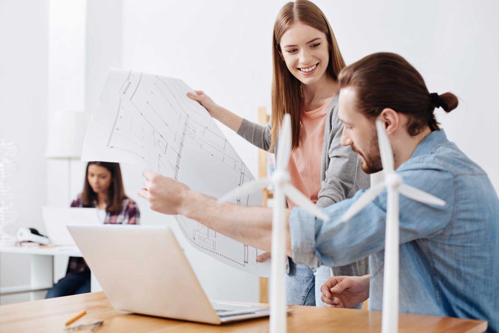 A man and woman discuss plans while reviewing a model of wind turbines, with a laptop and another person working in the background.