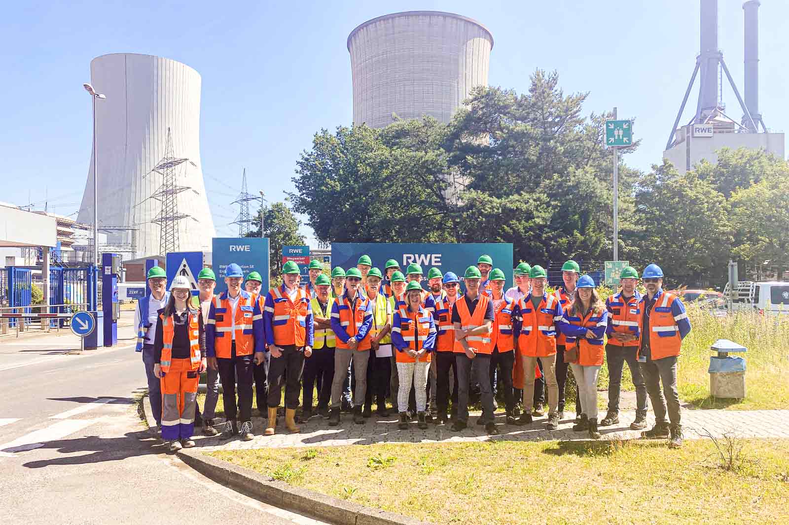 A group of workers in orange vests and hard hats stands in front of a power plant with cooling towers in the background.