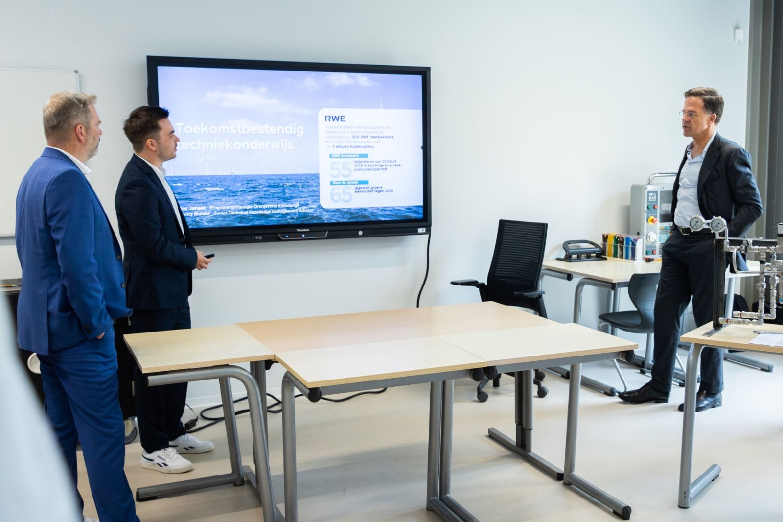 Three men discussing technology in a modern classroom, with a large screen displaying educational content and ocean imagery.