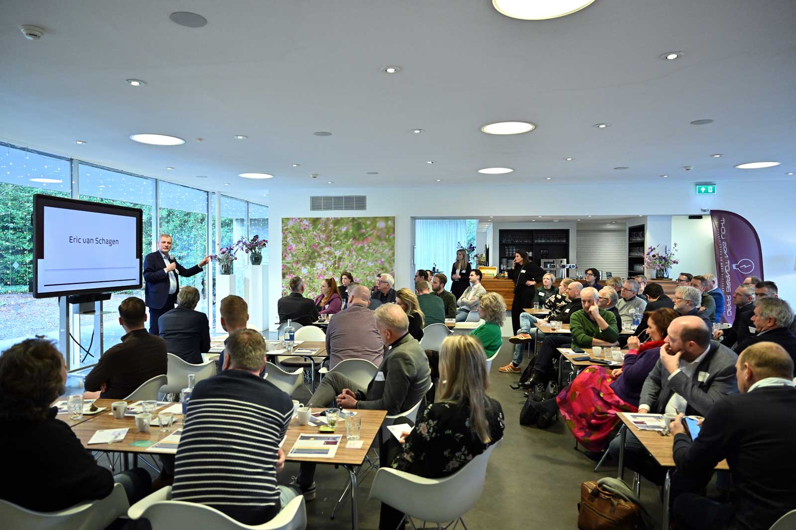 A conference with attendees seated at tables, focusing on a presenter named Eric van Schagen displaying a presentation.