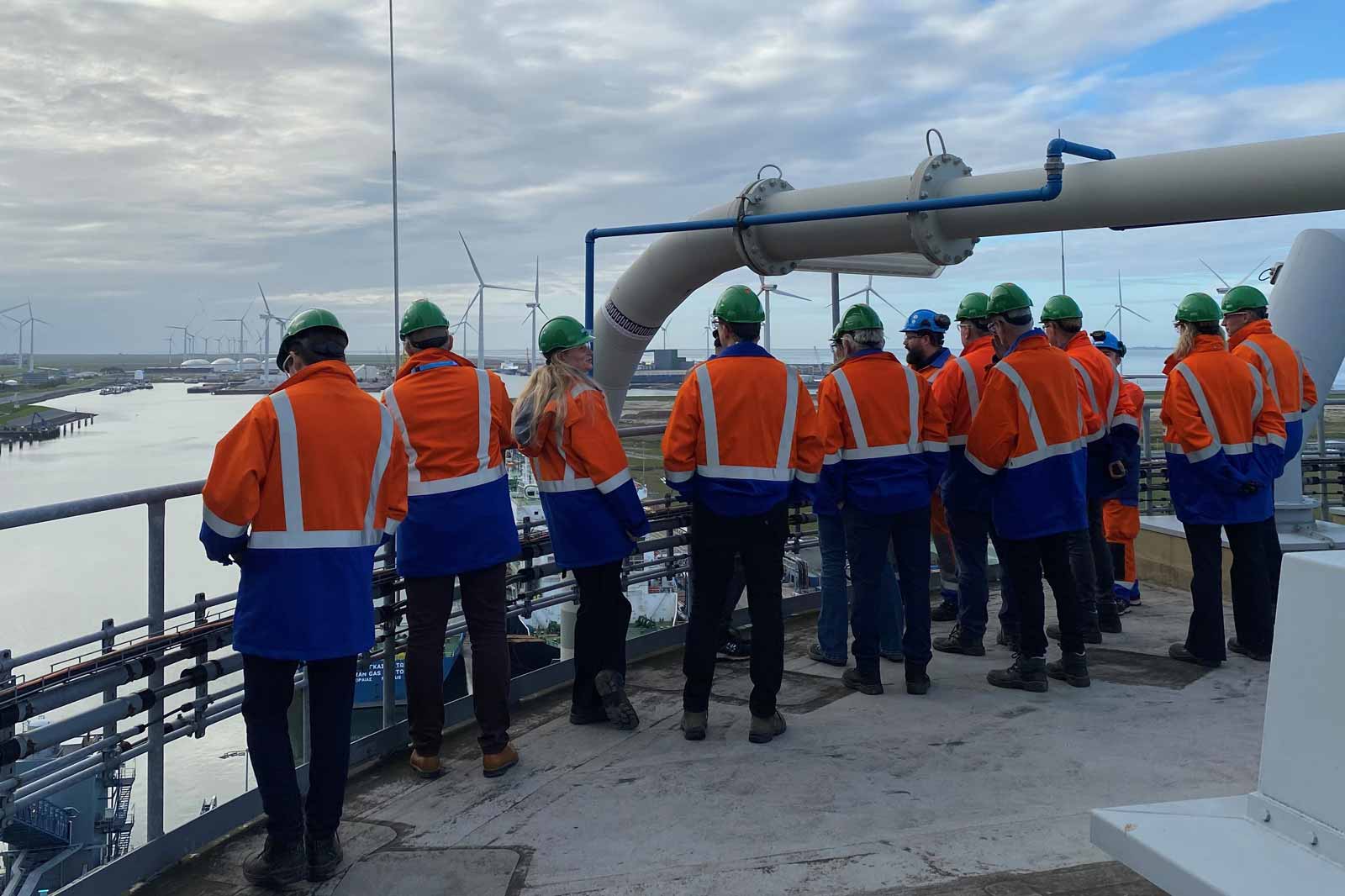 A group of workers in orange and blue safety gear overlooking a river with wind turbines in the background.