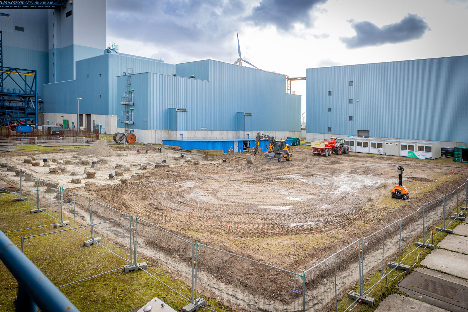 Een bouwplaats met machines en een omheind gebied, omgeven door grote blauwe gebouwen en een bewolkte lucht.