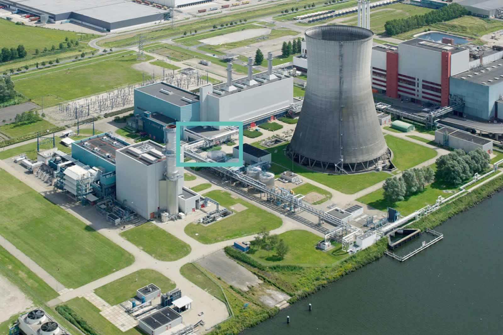 Luchtfoto van een energiecentrale met een koeltoren, industriële gebouwen en groene gebieden langs een waterlichaam.