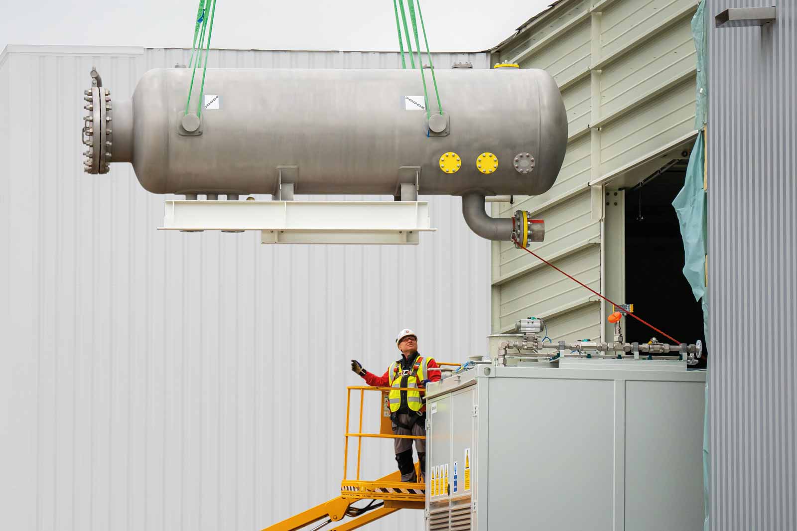 Een werknemer in opvallende veiligheidskleding bedient een kraan die een grote metalen tank in een gebouw hijst, met zichtbare veiligheidsvoorzieningen.
