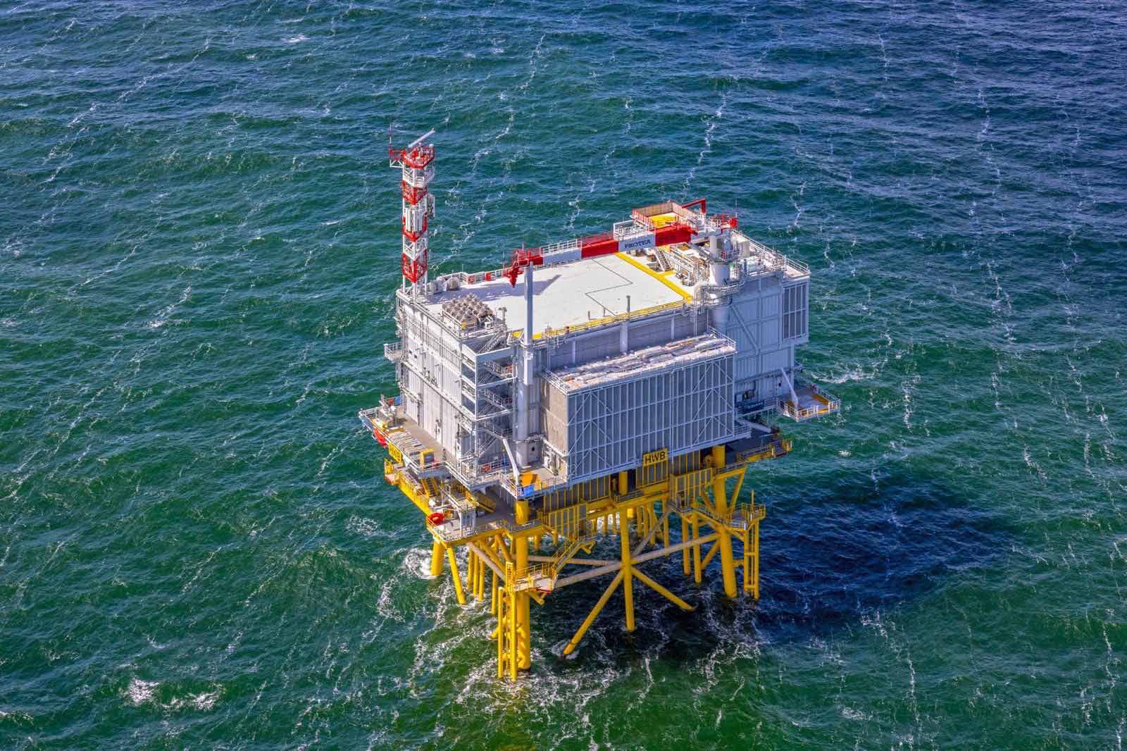 Een offshore platform staat boven groene golven, met rode en witte structuren en gele steunpijlers.