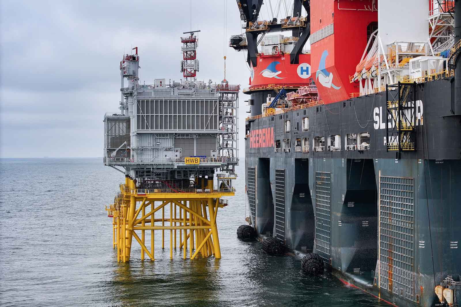 Een groot offshore platform met een gele ondersteuningsstructuur wordt onderhouden door een schip genaamd Sleipnir in een grijze oceaanomgeving.
