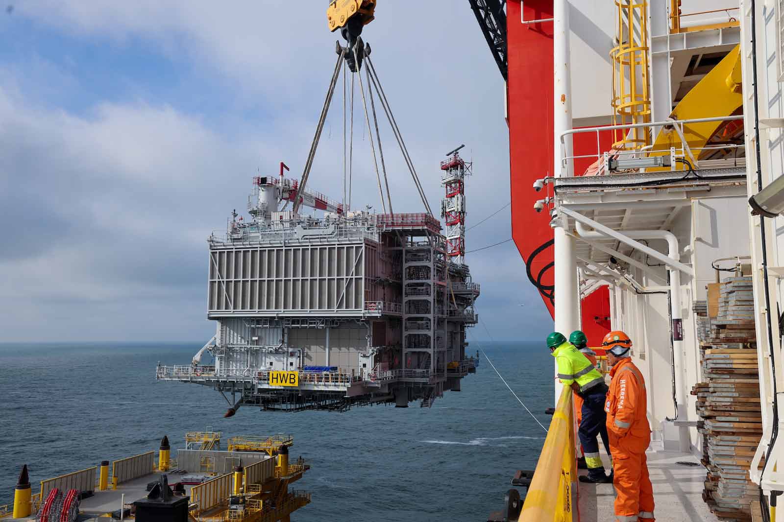 Een kraan tilt een groot offshore platformmodule boven de zee, met werknemers in veiligheidsgerei die vanaf een nabijgelegen schip toekijken.