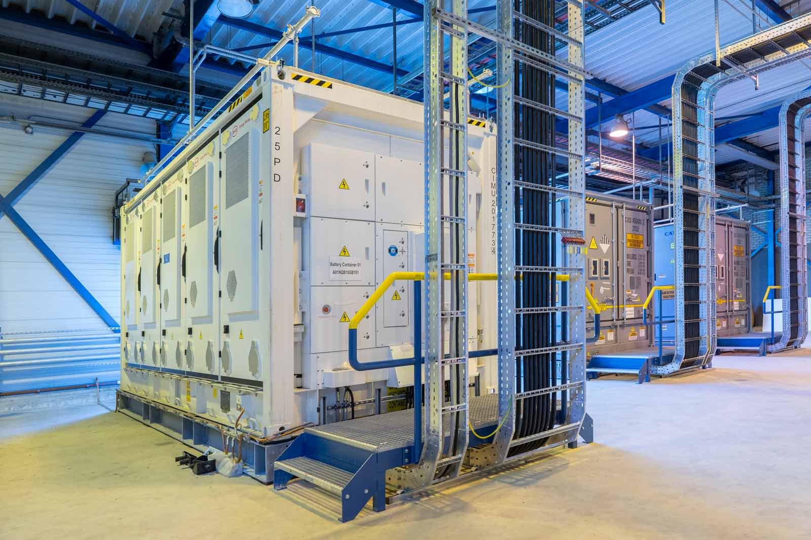 Een moderne batterijopslagfaciliteit met meerdere witte containers en gele veiligheidshekken in een goed verlichte industriële ruimte.