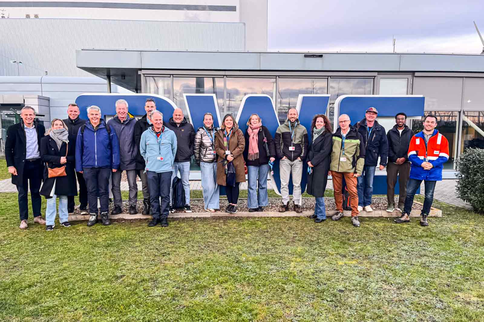 Een groep mensen staat samen voor een blauw bord, met een grasgebied en een modern gebouw op de achtergrond.