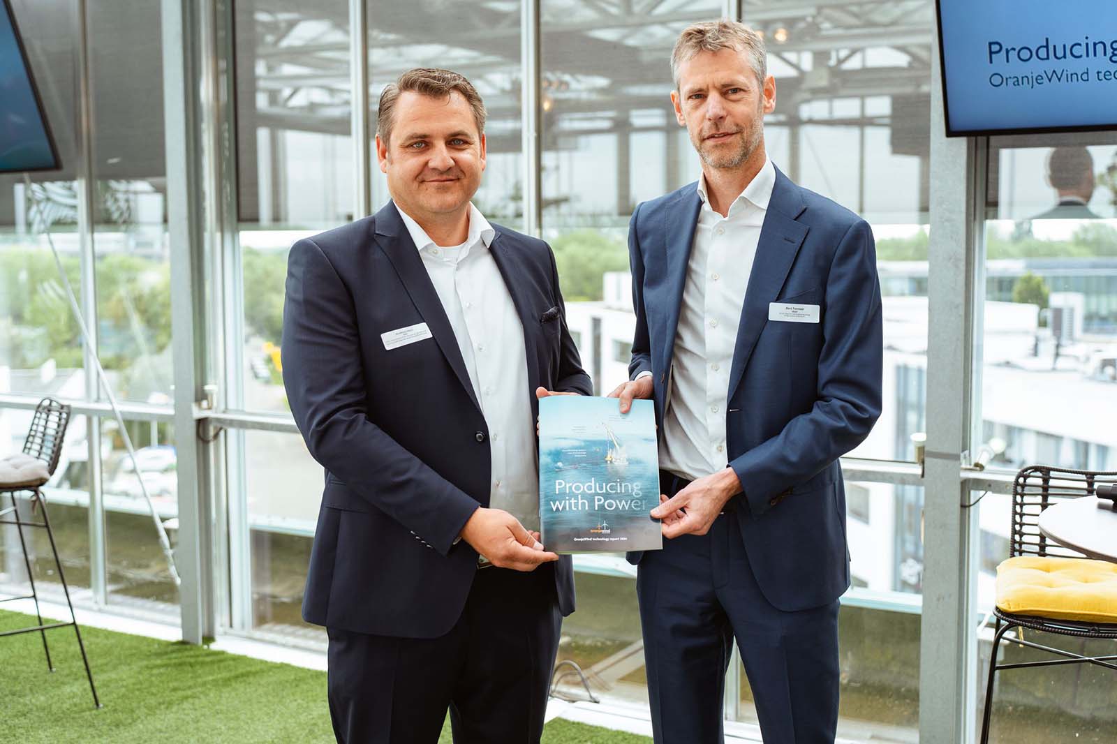 Twee mannen in zakelijke pakken staan binnen en houden een brochure vast met als titel "Producing with Power" met een modern kantoor op de achtergrond.