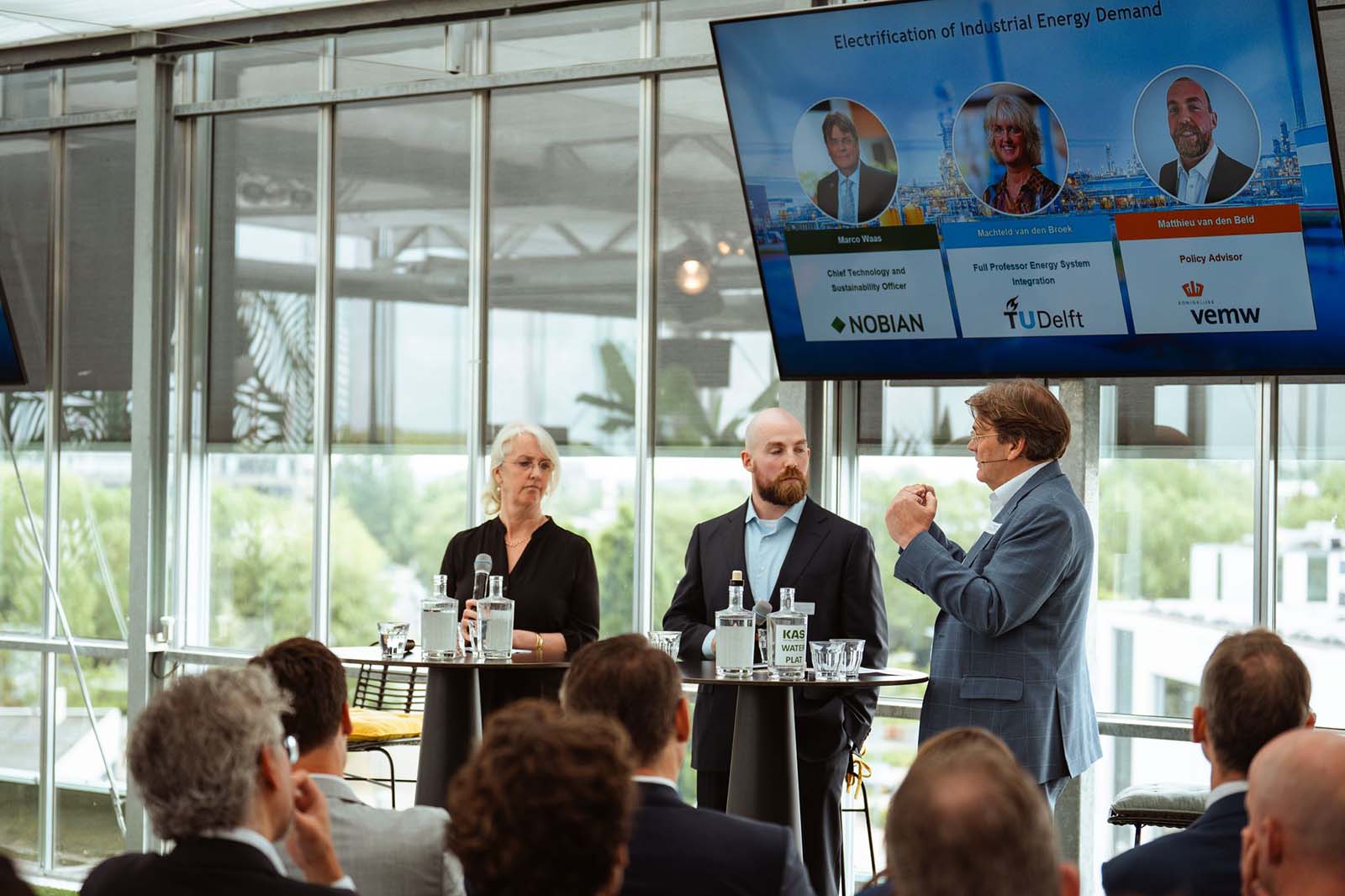 Drie sprekers staan achter twee hoge tafels met waterglazen en flessen, en geven een presentatie over industriÃ«le energievraag aan een publiek.