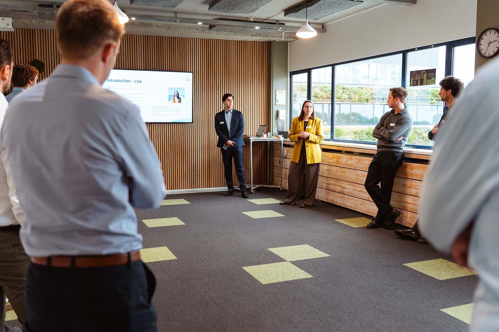 A group of professionals standing in a modern office space with wooden panel walls and large windows, listening to a presentation.