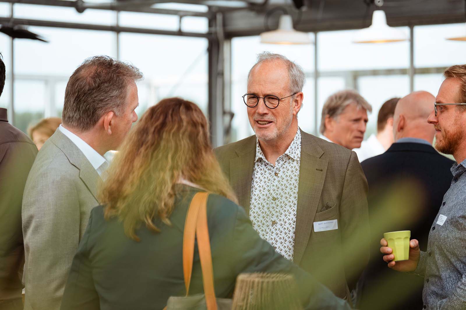 Een groep mensen die met elkaar praat tijdens een professioneel binnen evenement, waarbij iemand een groene beker vasthoudt.