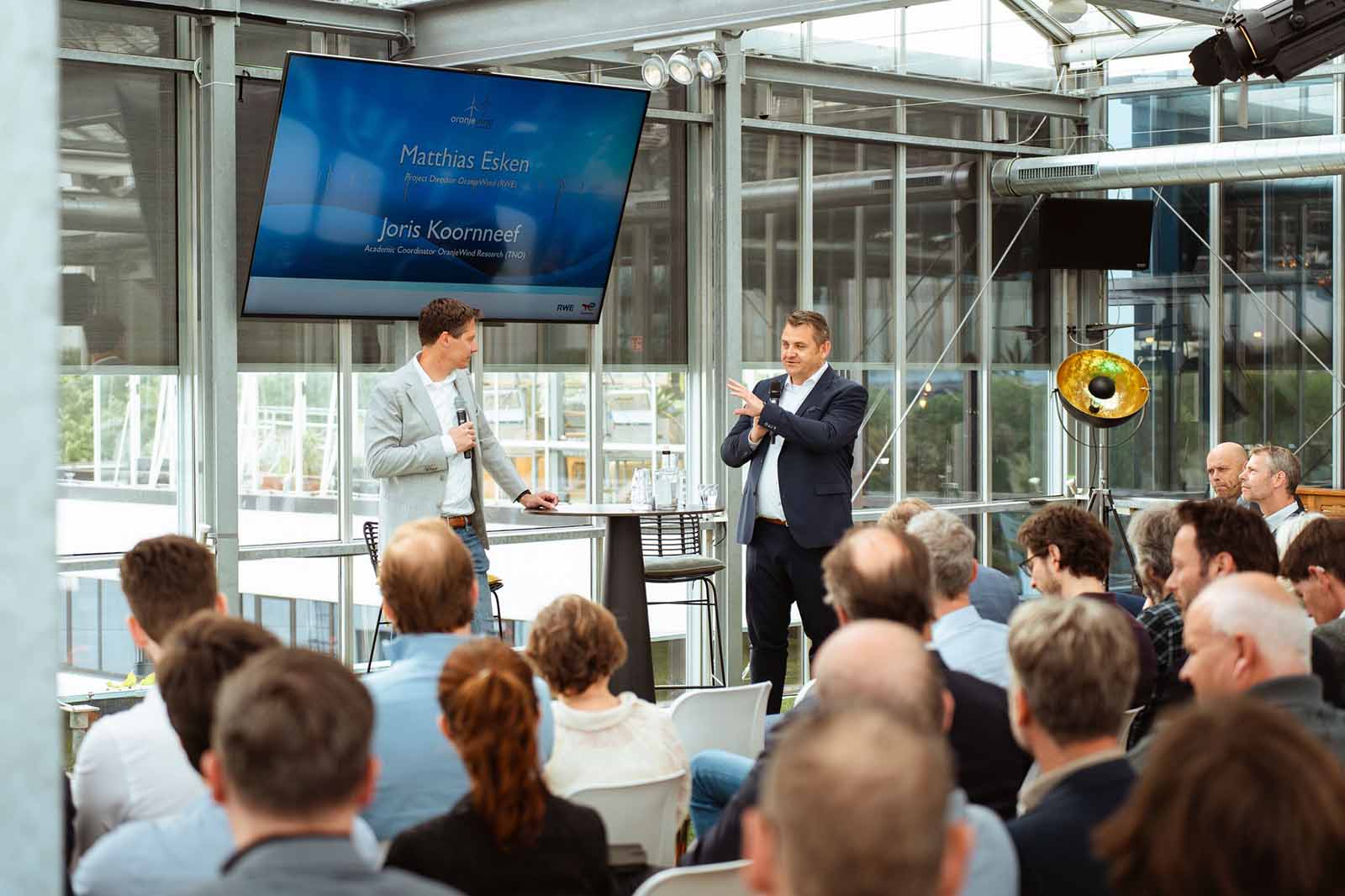 Two men are presenting in front of an audience inside a glass-walled room with a large screen displaying their names and titles.