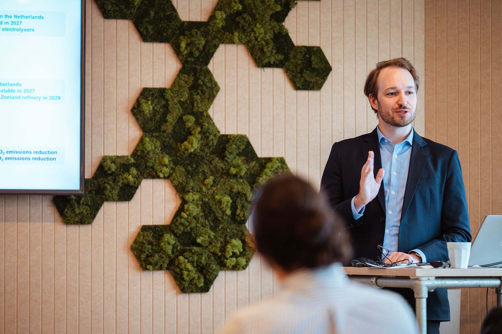 Een man in pak geeft een presentatie in een moderne kamer met hexagonale mosdecoraties aan de muur en een laptop op het bureau.