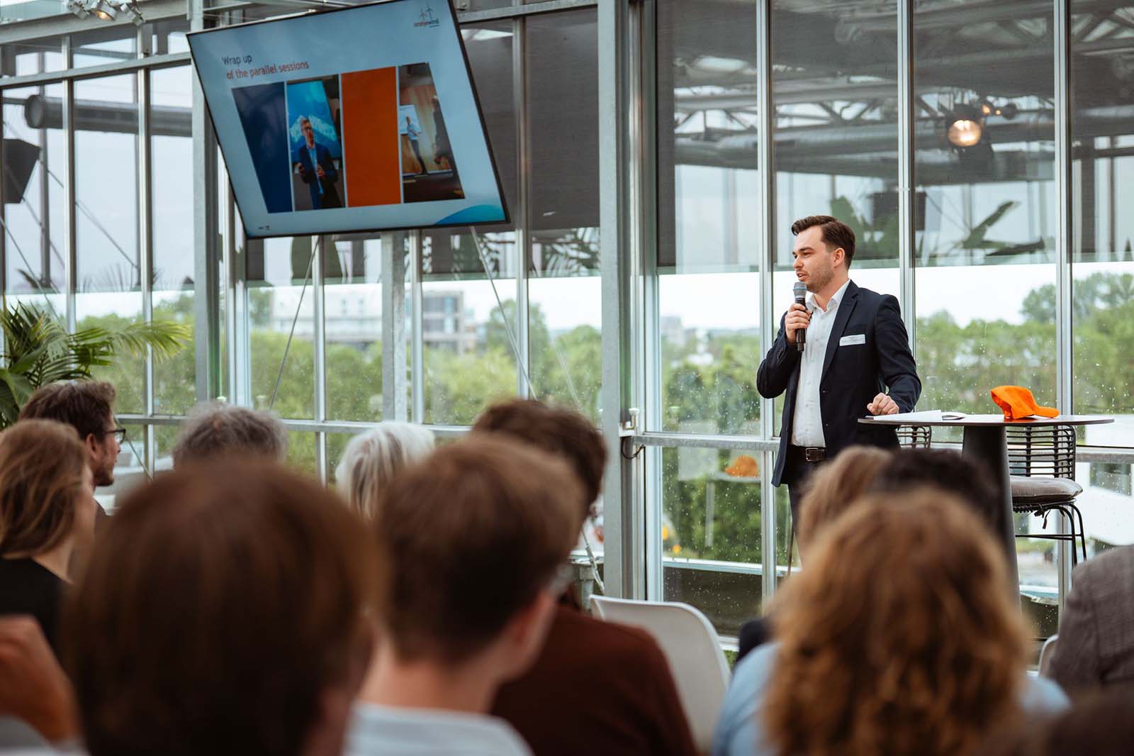 A man in a suit is speaking into a microphone at a modern conference room with large windows and a screen showing a presentation.