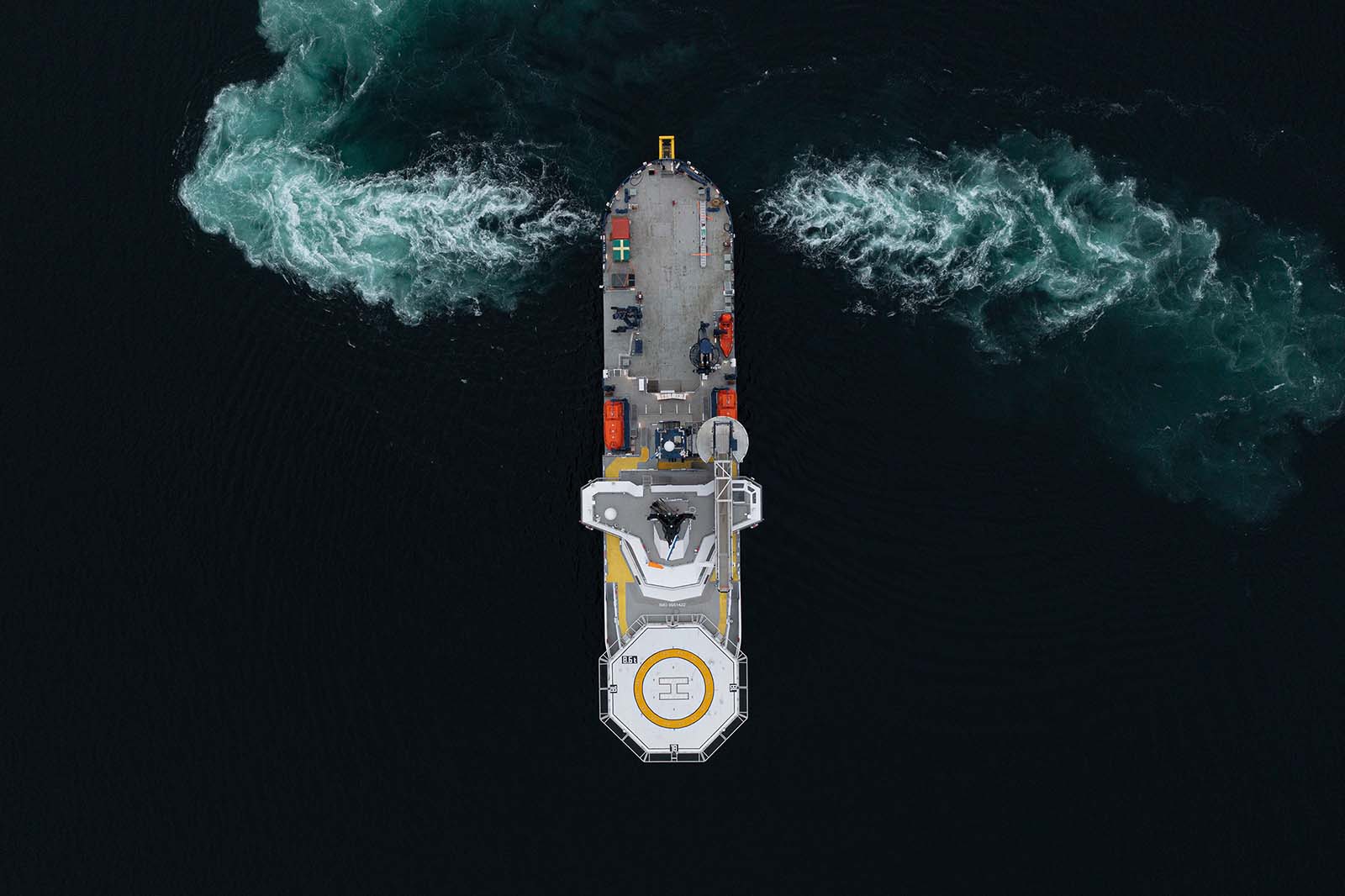 Luchtfoto van een schip in donker water met zichtbare witte waterstromen aan beide zijden, met een helikopterplatform en oranje reddingsboten.