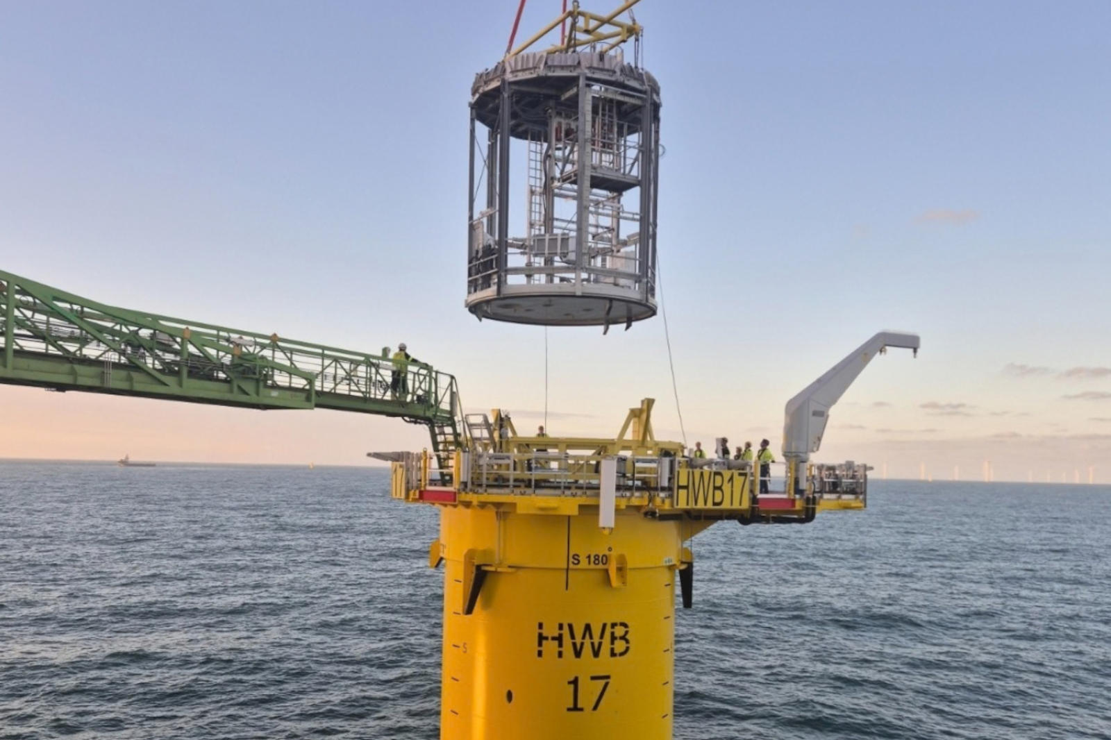 A yellow offshore platform tower labelled HWB 17 with workers and a suspended metal cage above it over the sea.
