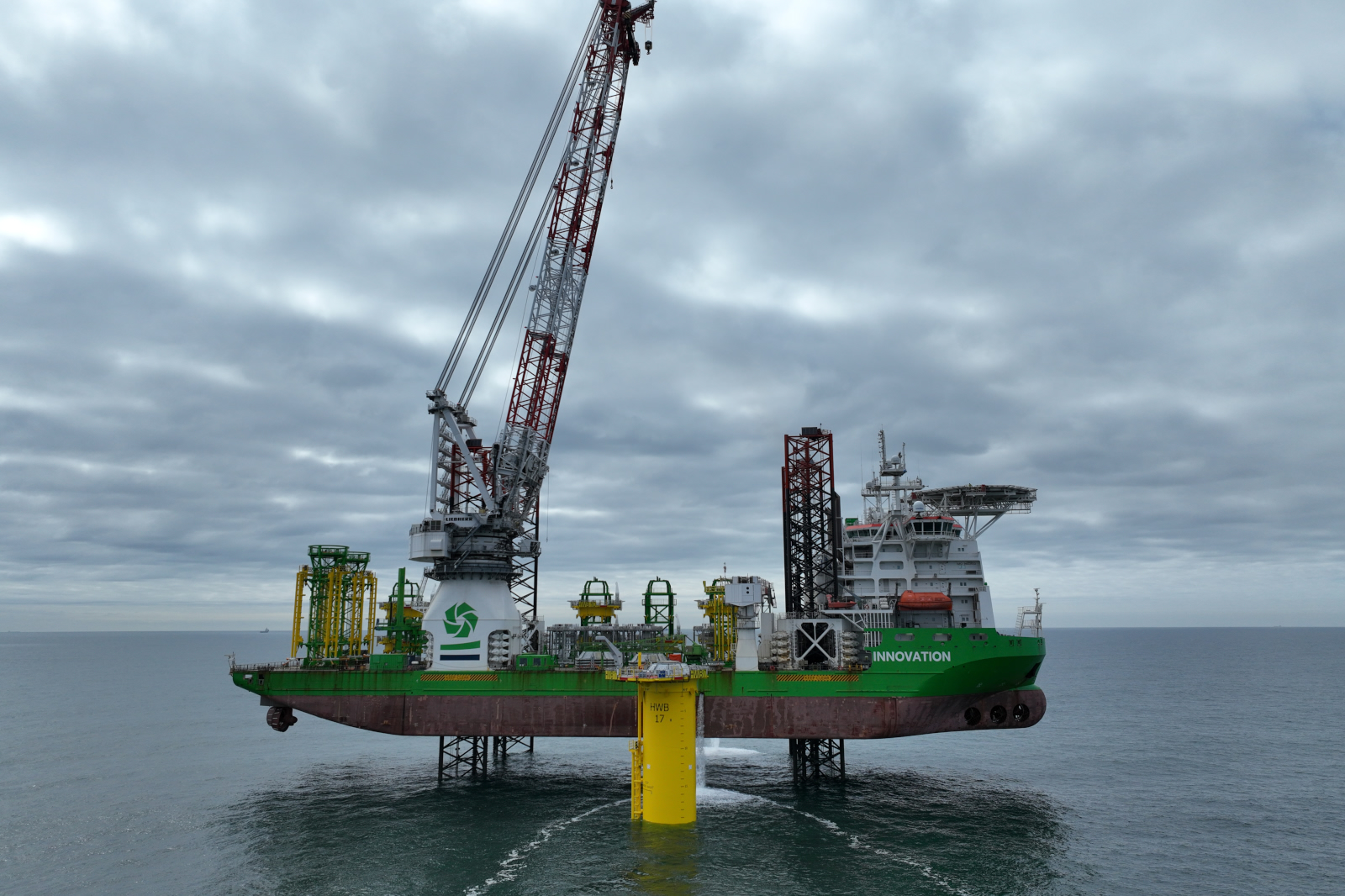 A green and white offshore jack-up vessel named Innovation with cranes is elevated above the sea on yellow legs under a cloudy sky.
