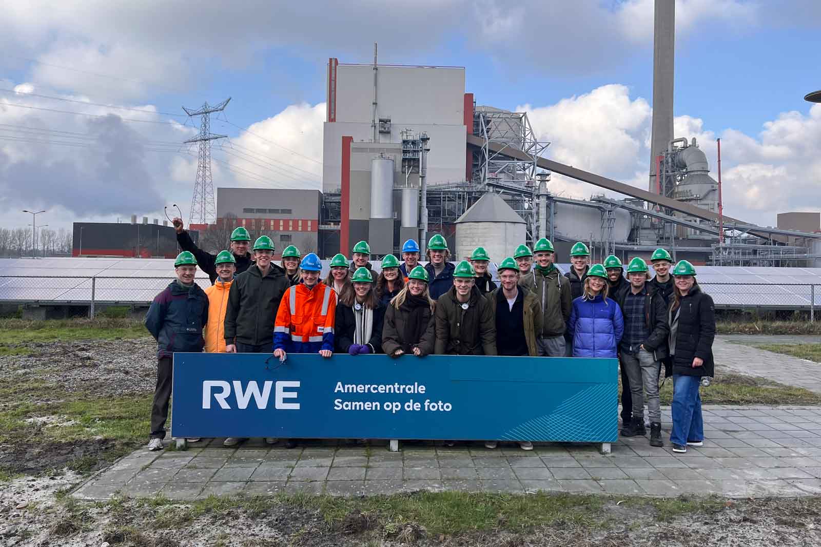 A group of individuals wearing green hard hats poses in front of an industrial facility with solar panels and electric towers.