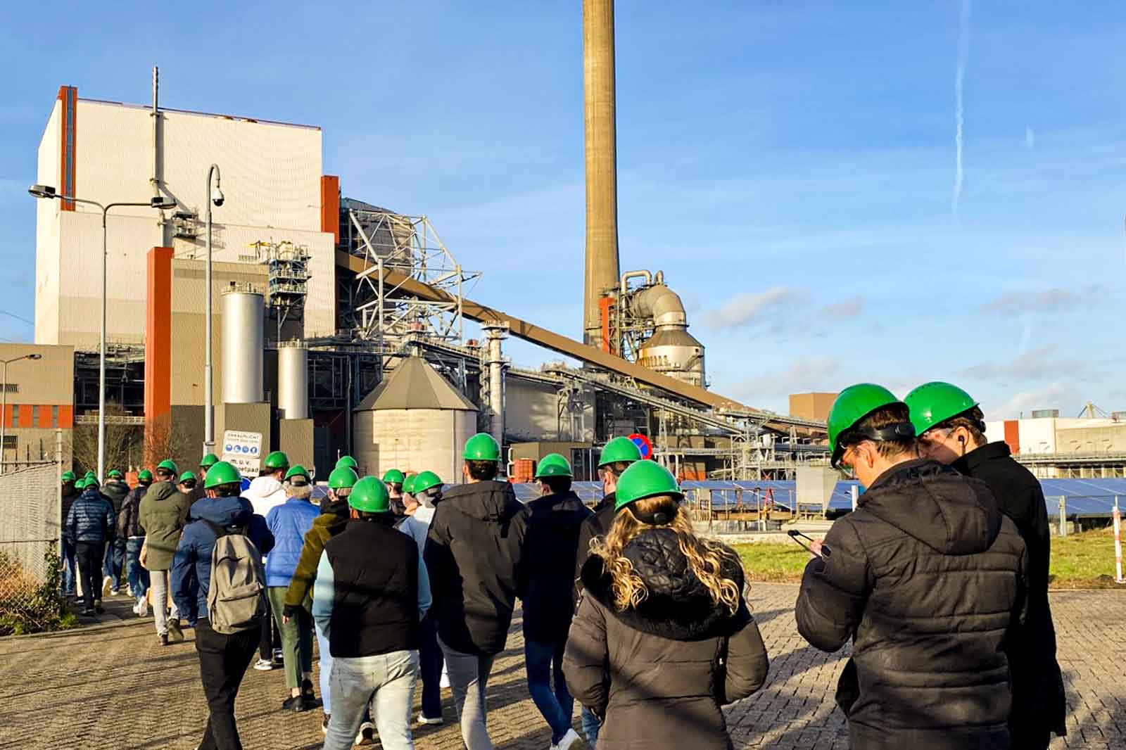 Een groep mensen met groene helmen loopt naar een industriële faciliteit onder een blauwe lucht.