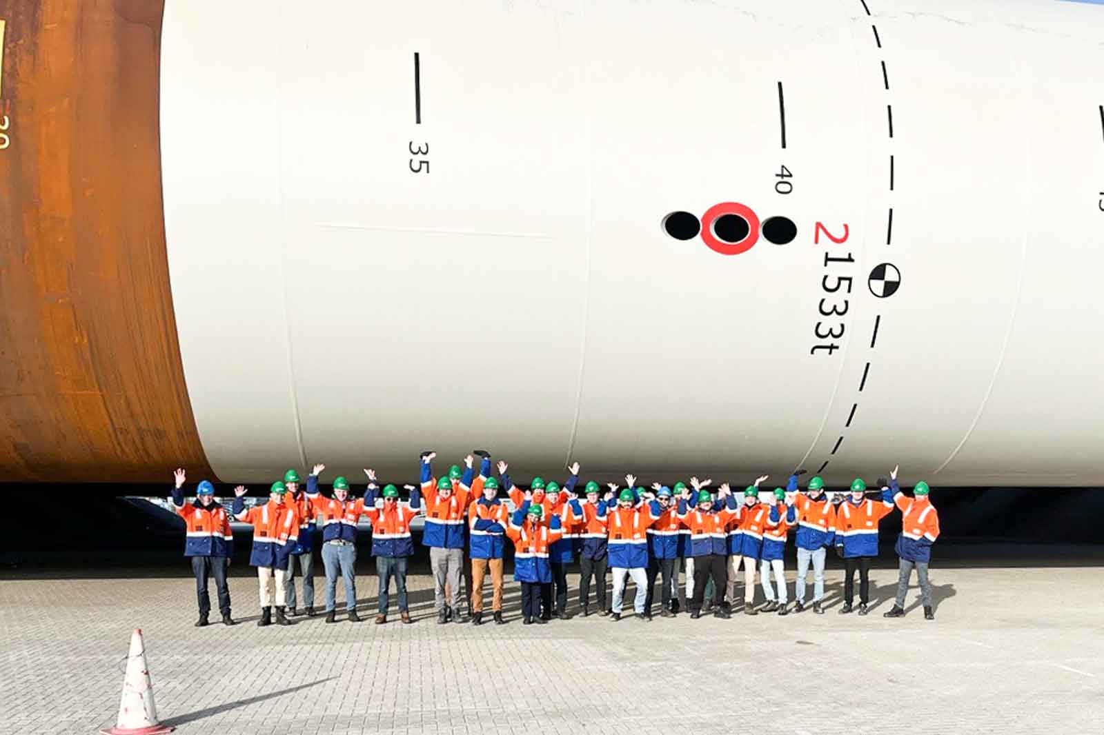 Een groep werknemers in oranje vesten en groene helmen staat onder een grote cilindrische structuur en zwaait enthousiast.