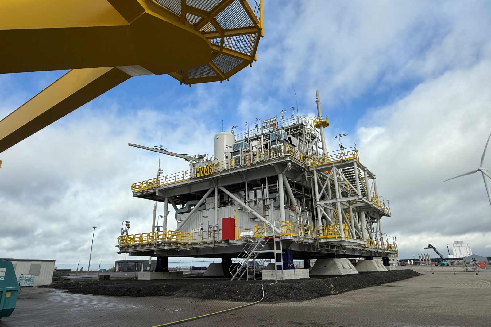 Een groot offshore platform genaamd HNA6, met gele relingen en meerdere structuren, gelegen op een bewolkte dag aan de kust.