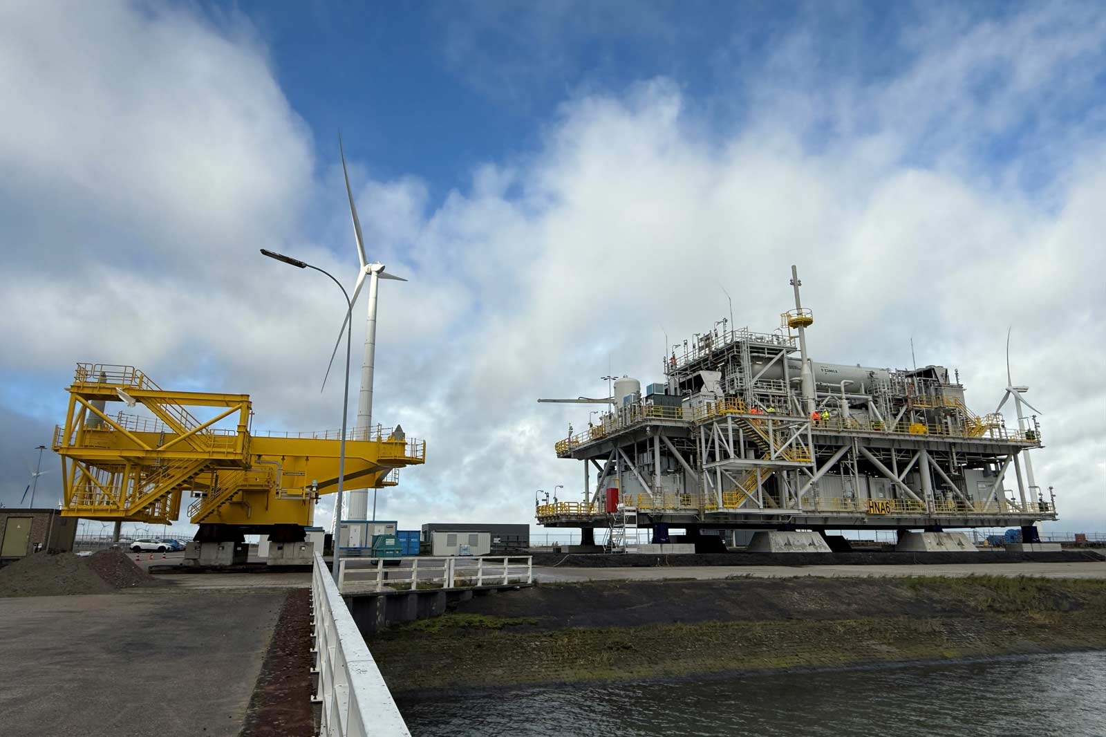 A yellow offshore platform sits alongside an industrial structure with wind turbines against a partly cloudy sky.