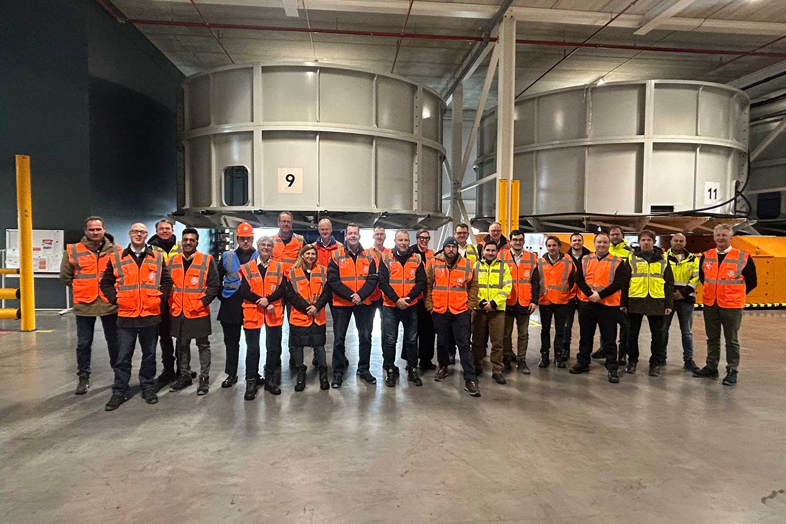 A group of people in safety vests poses inside a large industrial space, with equipment in the background.