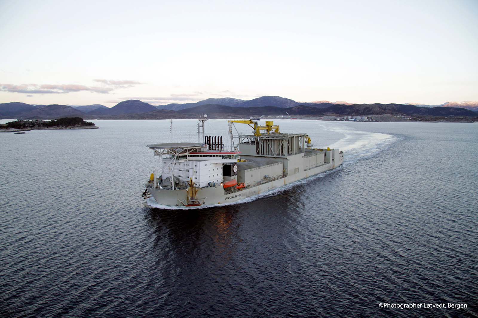 The ship named Simon Stevin sails through calm waters near mountainous coastline during early evening.