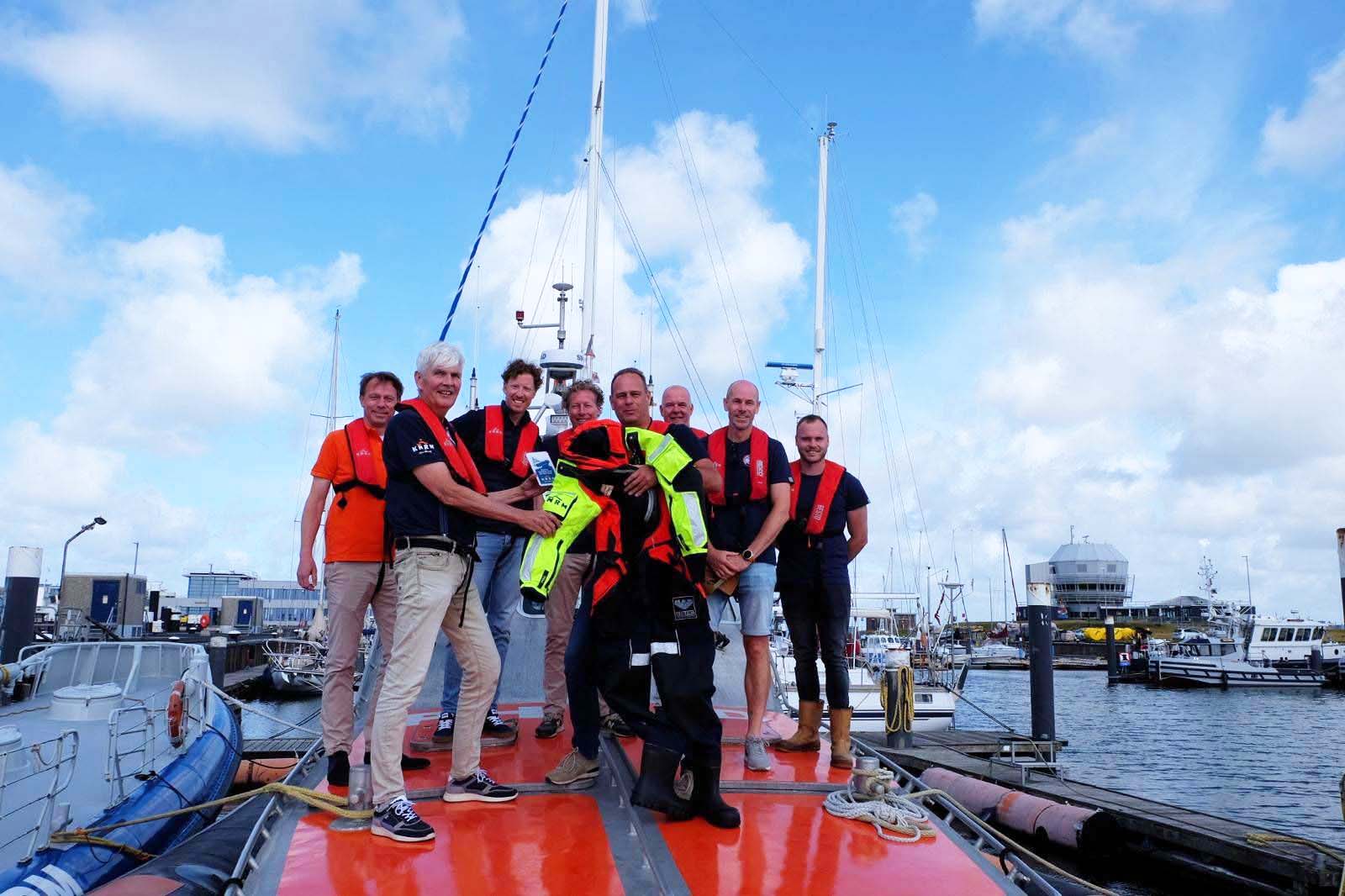 Een groep van acht mannen met reddingsvesten staat op een aangemeerde boot en houdt een felgeel en zwart reddingspak vast onder een licht bewolkte hemel.