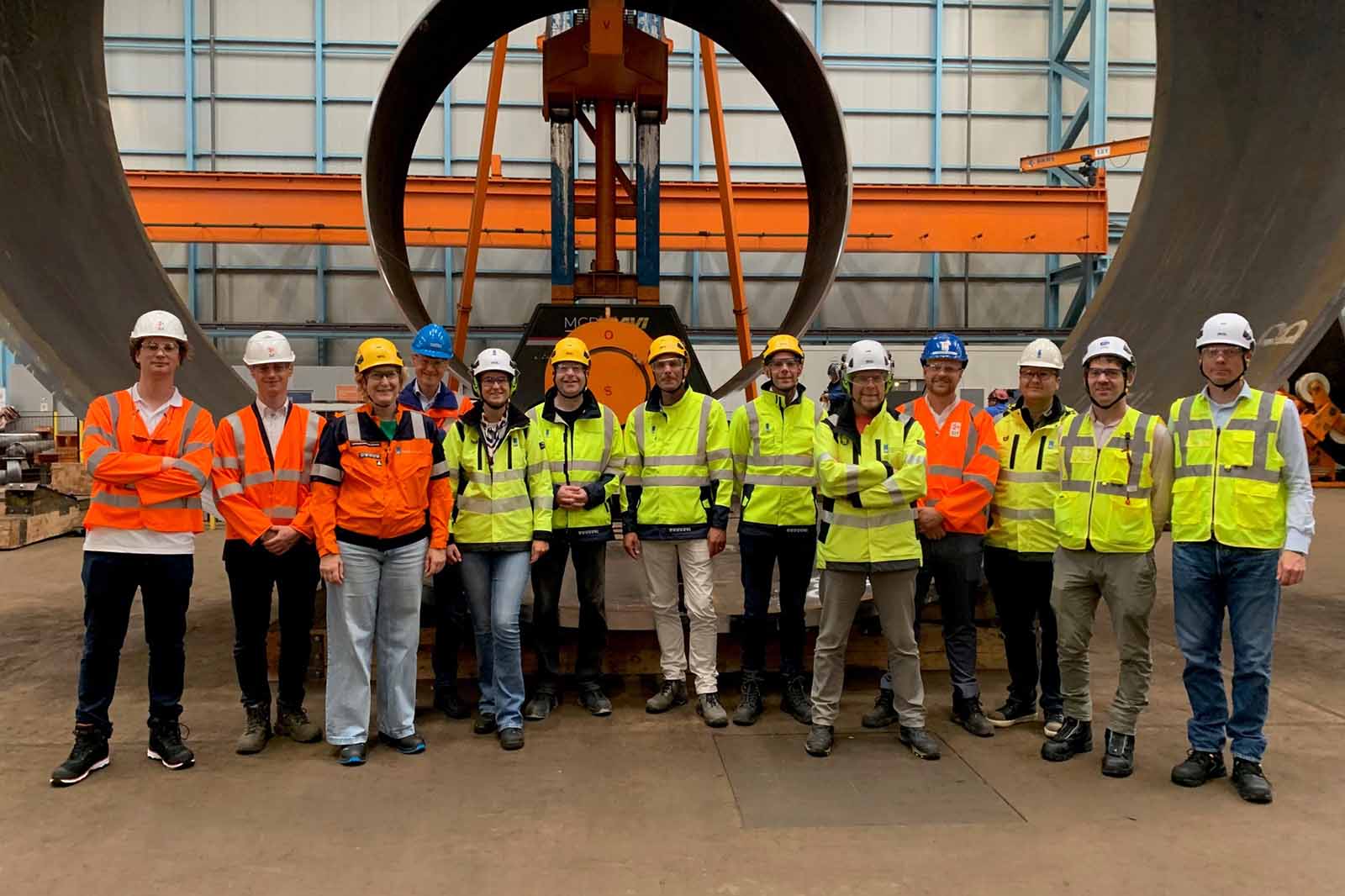 A group of workers wearing safety helmets and high-visibility jackets pose inside an industrial facility with large metal pipes.