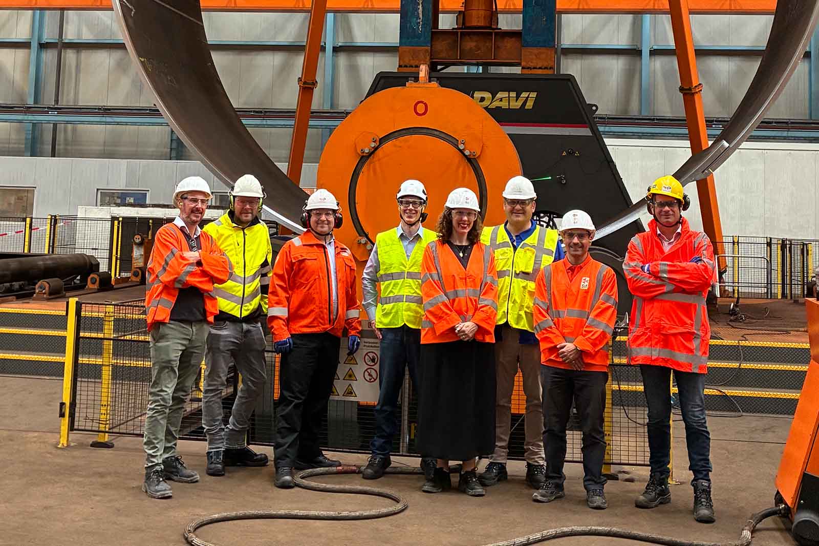 Eight workers wearing high-visibility jackets and helmets stand in front of large industrial machinery inside a factory.