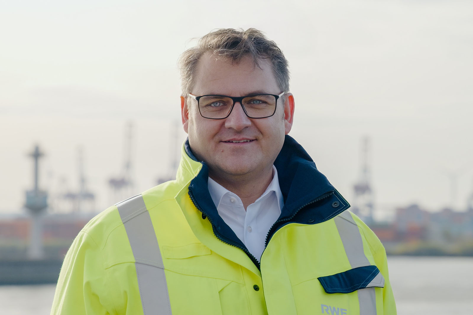 Man met bril en geel veiligheidsjack staat buiten met industriële kranen op de achtergrond.