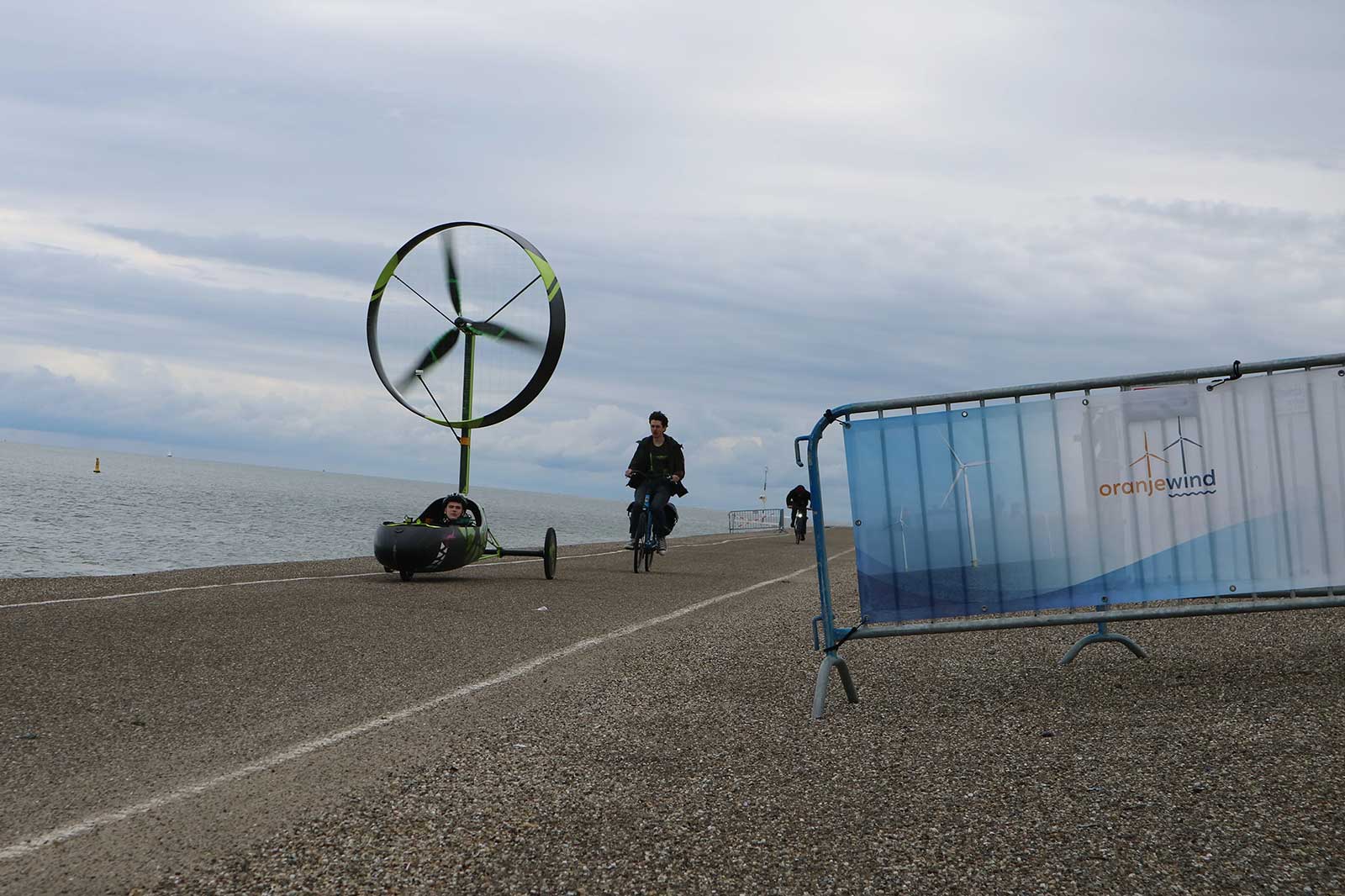 A small wind-powered vehicle and a cyclist ride on a coastal pathway under a cloudy sky.