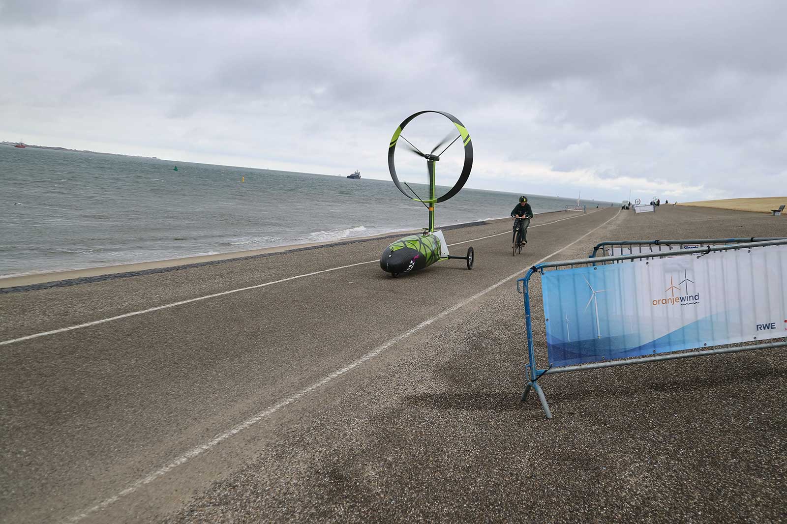 A cyclist rides near a small green wind-powered vehicle on a coastal road by the sea under a cloudy sky.