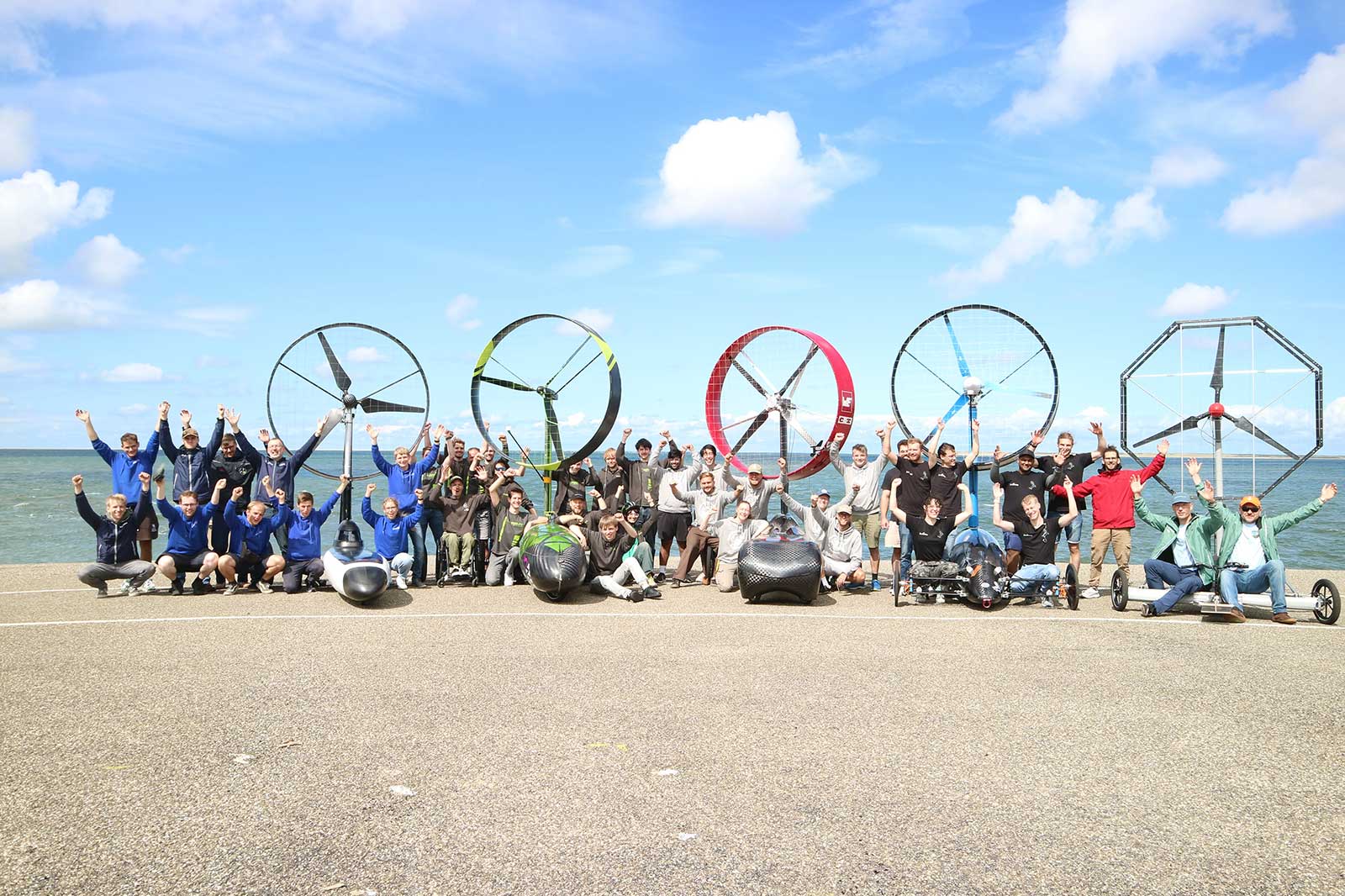 A large group of people in front of the sea cheer while sitting and standing behind human-powered vehicles with large propellers.