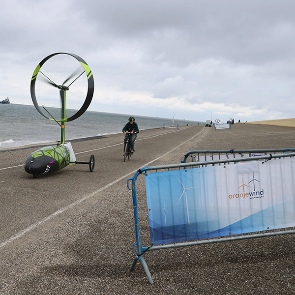 Een persoon fietst op een kustpromenade naast een windgedreven driewieler met een groot cirkelvormig blad onder een bewolkte lucht.